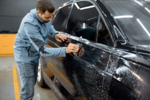 Car service worker sticking anti-gravel film on a car body with scrapper at the detailing vehicle workshop. Concept of car body protection with special films