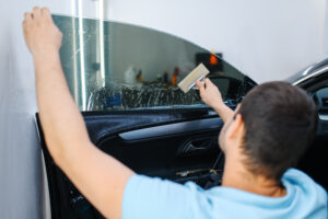 Male worker with squeegee installs car tinting, tuning service. Mechanic applying vinyl tint on vehicle window in garage, tinted automobile glass