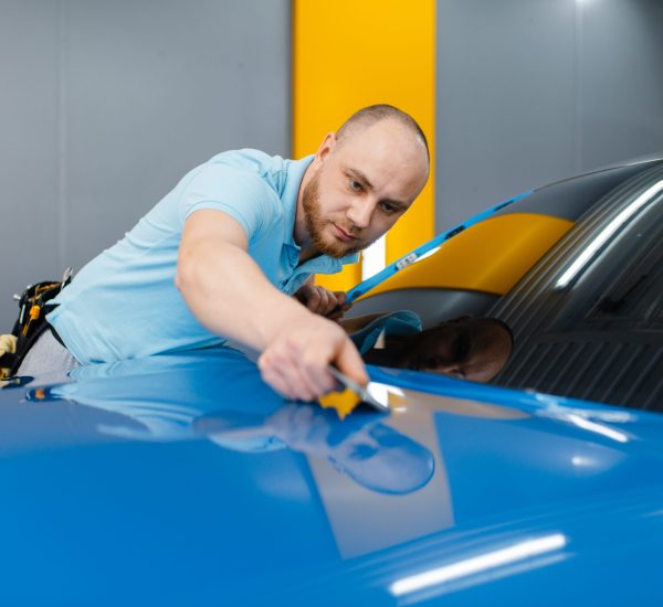 Male car wrapper with squeegee installs protective vinyl foil or film on hood. Worker makes auto detailing. Automobile paint protection coating, professional tuning