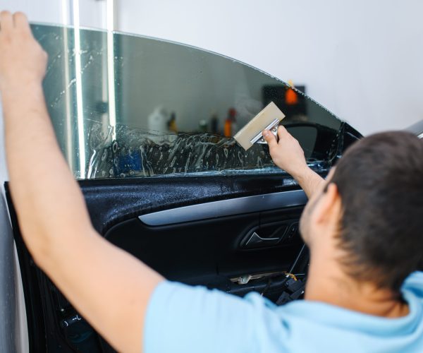 Male worker with squeegee installs car tinting, tuning service. Mechanic applying vinyl tint on vehicle window in garage, tinted automobile glass