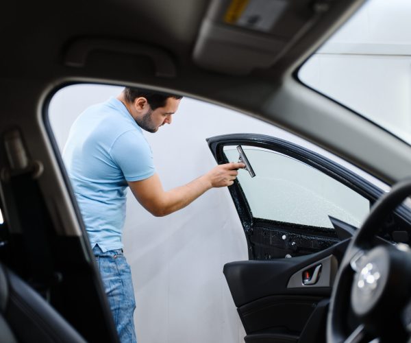 Male wrapper installs car tinting, tuning service. Worker applying vinyl tint on vehicle window in garage, tinted automobile glass